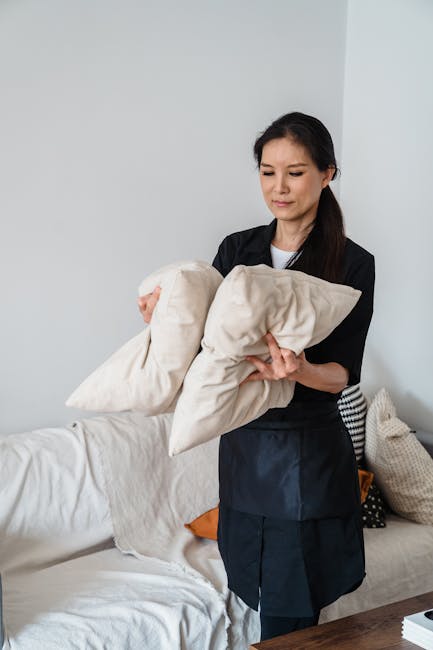 A professional cleaner from Carpet Cleaners Knightsbridge is depicted indoors, standing beside a light-colored sofa in a well-lit living room. She is holding a beige pillow and a matching cushion, appearing to be preparing for a surface cleaning or upholstery revitalisation. The sofa's fabric looks clean and fresh, with a tidy arrangement of cushions, including a patterned and an orange cushion. The background features a plain white wall and a wooden table, emphasizing a neat, domestic setting. The room's lighting is bright, highlighting the clean and well-maintained condition of the furnishings, consistent with deep cleaning or upholstery care services offered by Carpet Cleaners Knightsbridge as part of a comprehensive sanitisation and domestic cleaning process.