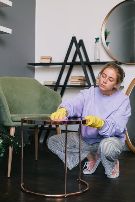 A professional cleaner from Carpet Cleaners Knightsbridge is depicted indoors, standing beside a light-colored sofa in a well-lit living room. She is holding a beige pillow and a matching cushion, appearing to be preparing for a surface cleaning or upholstery revitalisation. The sofa's fabric looks clean and fresh, with a tidy arrangement of cushions, including a patterned and an orange cushion. The background features a plain white wall and a wooden table, emphasizing a neat, domestic setting. The room's lighting is bright, highlighting the clean and well-maintained condition of the furnishings, consistent with deep cleaning or upholstery care services offered by Carpet Cleaners Knightsbridge as part of a comprehensive sanitisation and domestic cleaning process.