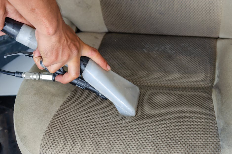A professional cleaner from Carpet Cleaners Knightsbridge, dressed in a black uniform, is preparing to deep clean a beige upholstered sofa with a white cloth in a well-lit living room. The room featur