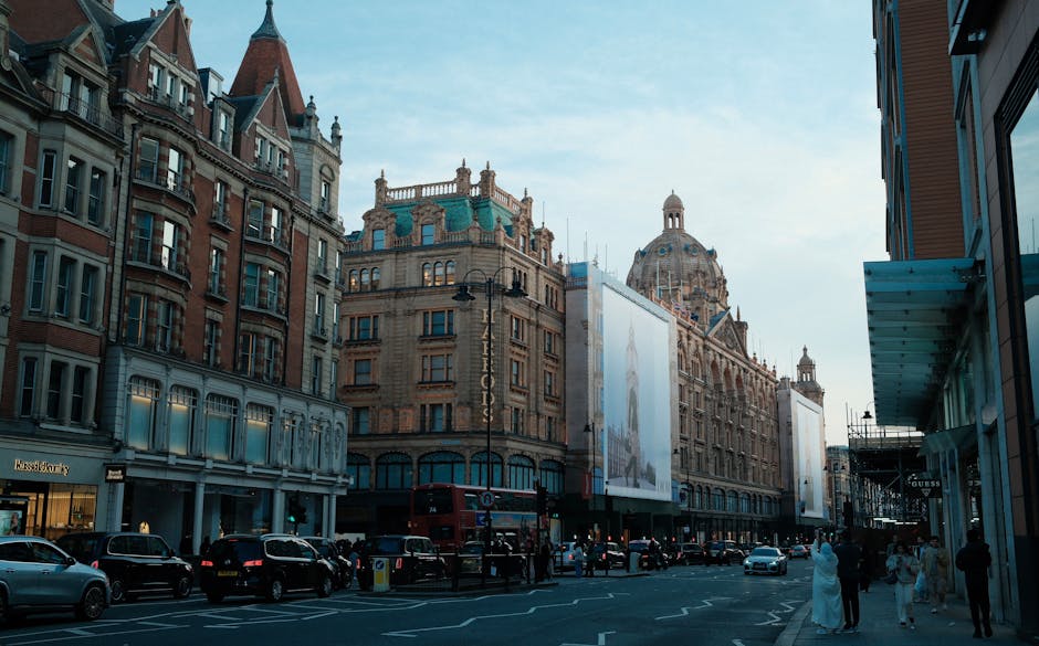 A busy street scene in Knightsbridge with historic and modern buildings lining the road, including luxury storefronts and a large advertising billboard mounted on a historic building. Pedestrians are walking along the sidewalk, some talking, others window shopping, while cars, including black and white models, are parked or driving past. The sky is clear with a few clouds, and the street is well-lit, reflecting a vibrant commercial area. COVID-19 safety measures like social distancing are subtly observed in the scene, with some people wearing face masks. Carpet Cleaners Knightsbridge, a professional cleaning company, provides surface cleaning and deep cleaning services in such areas to maintain hygiene and cleanliness of commercial spaces.