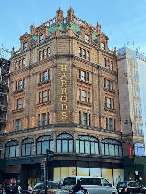 Exterior view of the historic Harrods building in Knightsbridge, showcasing ornate stone architecture with decorative turrets and large ground-floor windows. The building's facade features a prominent vertical yellow sign spelling 'HARRORDS,' and the street below includes parked cars, a cyclist, and pedestrians. Bright daylight illuminates the facade, emphasizing the intricate detailing of the architectural features. Carpet Cleaners Knightsbridge specializes in surface cleaning and deep cleaning services, ensuring the maintenance and hygienic upkeep of such commercial properties, along with residential spaces. This image highlights the importance of professional sanitisation and cleanliness standards in busy urban environments.