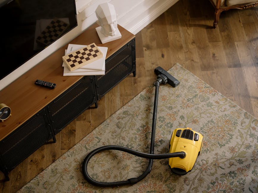 A yellow vacuum cleaner with a black hose attachment is placed on a beige floral patterned area rug in a living room. The room features wooden flooring and a black metal and wood media console against the wall, topped with a white sculpture, a closed book with a chessboard pattern, a stack of papers, and a black remote control. The lighting is natural, illuminating the space evenly. Carpet Cleaners Knightsbridge specializes in surface cleaning and deep cleaning services, ensuring hygienic and well-maintained interiors, as highlighted in their tips to avoid common cleaning mistakes.