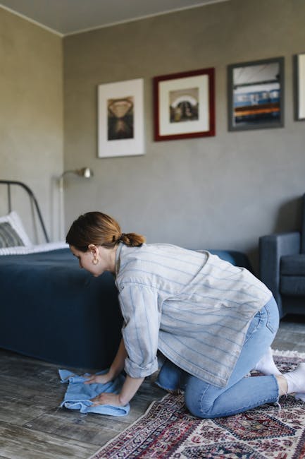 A woman with brown hair tied back, dressed in a light blue striped shirt and jeans, is kneeling on a patterned rug and cleaning a wooden floor with a blue microfiber cloth. The room features a grey wall decorated with three framed art prints above a bed with a metal headboard, and a blue upholstered chair is partially visible in the background. The space is well-lit with natural light, highlighting the cleanliness and tidy arrangement, emphasizing surface cleaning and domestic hygiene practices which are relevant to avoiding mistakes in carpet cleaning and maintaining overall cleanliness, as advised by Carpet Cleaners Knightsbridge.
