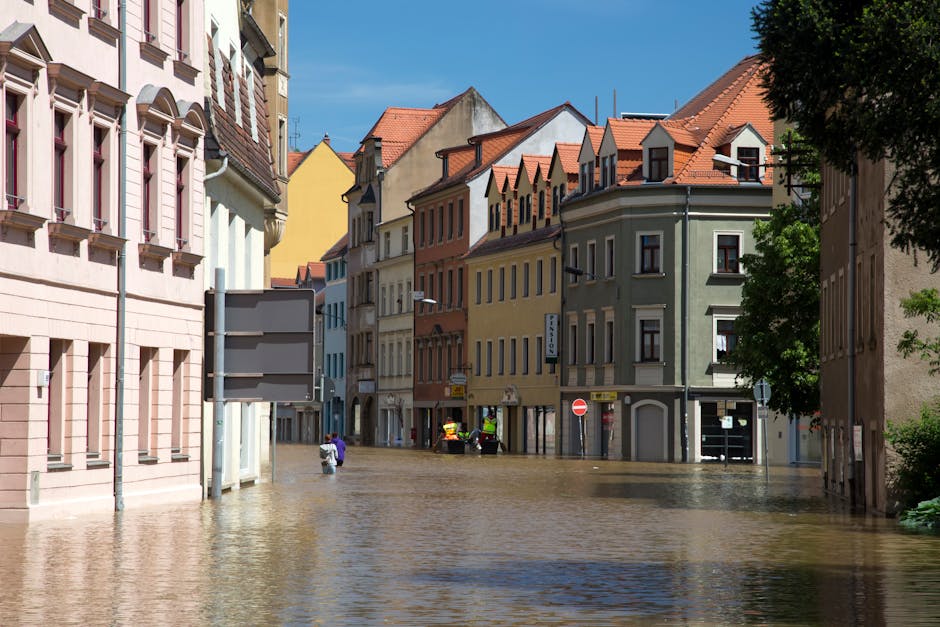 A street in Knightsbridge with multiple multi-story buildings featuring pastel and earth-toned facades, some with decorative window frames and small rooftop dormers. The street is heavily flooded with muddy water covering the entire road, reaching halfway up the doorways of ground-floor shops and residences. Emergency workers in high-visibility clothing are seen near the center, possibly assessing the situation. Bright daylight illuminates the scene, casting shadows from a large tree on the right side, while the sky above is clear blue. The scene captures the extent of flood damage in an urban residential area, highlighting the need for professional flood response and cleaning services, such as those offered by Carpet Cleaners Knightsbridge.
