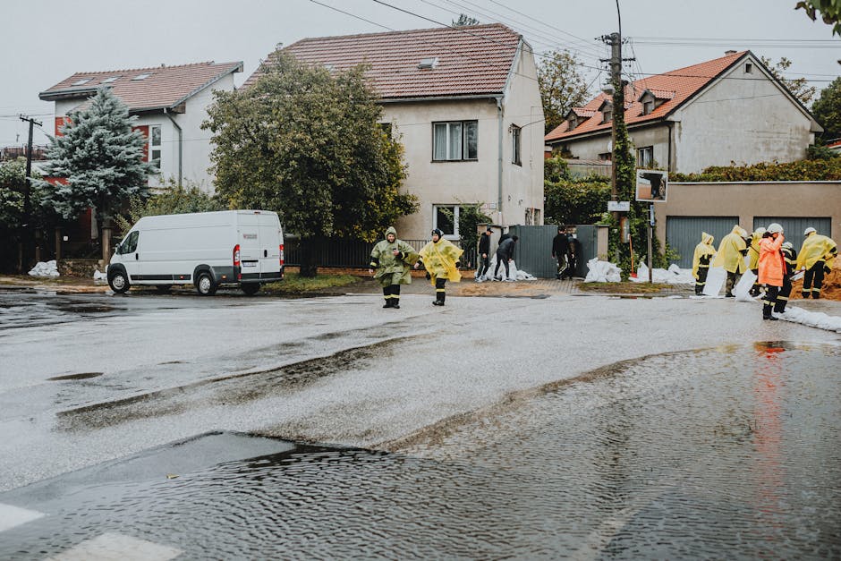 A residential street in Knightsbridge after flooding, with several emergency responders wearing yellow and orange raincoats walking across the wet, waterlogged pavement. Puddles cover the road surface, reflecting the overcast sky above. In the background, there are two-story houses with tiled roofs and windows, some obscured by trees and shrubs. A white van is parked at the curb, and debris is visible at the edges of the sidewalk. The scene suggests a coordinated effort to manage flood damage, with teams possibly involved in surface cleaning or sanitisation, highlighting the importance of professional emergency cleaning services provided by Carpet Cleaners Knightsbridge.