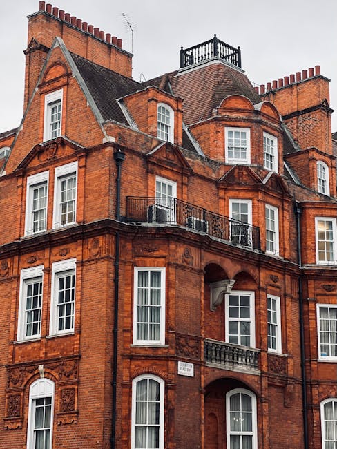 Image of a historic red brick residential building with multiple floors, featuring large white-framed sash windows, decorative stone moldings, and ornate cornices. The building has a gabled roof with slate shingles, a small balcony with a metal railing on the second floor, and a rooftop terrace enclosed by a black metal railing. The exterior appears clean and well-maintained, showcasing intricate architectural details typical of classical Victorian or Edwardian styles. The lighting is natural, indicating an overcast day. This setting emphasizes the importance of surface cleaning and preservation for maintaining the building's aesthetic appeal, consistent with the services offered by Carpet Cleaners Knightsbridge in the realm of deep cleaning and property maintenance.