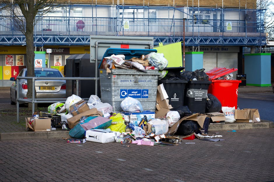 An overflowing outdoor waste collection area in a commercial or residential setting with multiple black, red, and grey rubbish bins filled with mixed waste, including cardboard boxes, plastic bags, paper, and packaging materials. The waste spills onto the surrounding brick pavement, with a grey recycling bin labeled 'mixed paper & card' among the overflowing refuse. Behind the bins, a metal railing and parked cars are visible on the street, with a building featuring multiple signage and a blue scaffolding structure in the background. The scene looks cluttered and unclean, emphasizing the need for proper waste disposal and regular cleaning highlighted by Carpet Cleaners Knightsbridge's services, especially in relation to waste rules for mattress and carpet disposal after cleaning in the RBKC area.