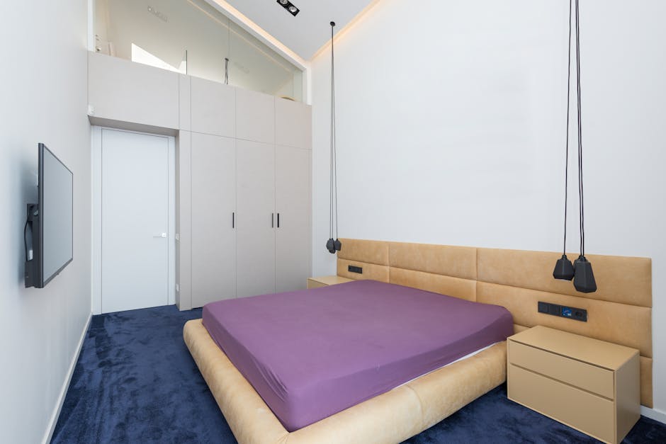 A modern bedroom featuring a beige upholstered bed with a purple mattress, flanked by two small beige nightstands, illuminated by two hanging black pendant lights. The room has white walls with a high ceiling and recessed lighting, a large built-in wardrobe with white doors on one side, and a flat-screen television mounted on the adjacent wall. The flooring is covered with a deep blue carpet, and the room appears clean and well-maintained, reflecting professional cleaning and hygiene standards typical of domestic cleaning services by Carpet Cleaners Knightsbridge, especially relevant to surface cleaning and sanitisation after mattress disposal guidelines.