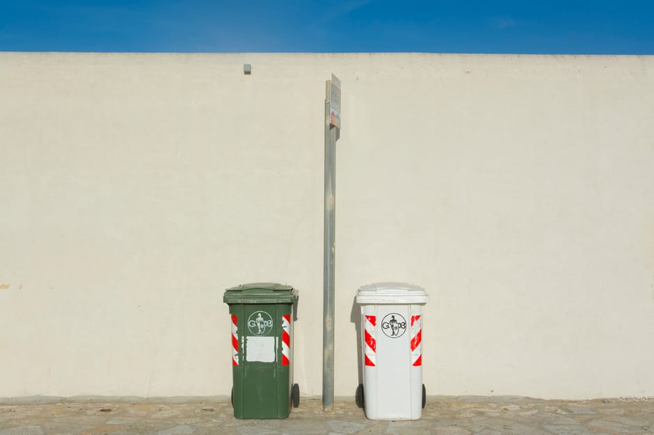 An overflowing outdoor waste collection area in a commercial or residential setting with multiple black, red, and grey rubbish bins filled with mixed waste, including cardboard boxes, plastic bags, paper, and packaging materials. The waste spills onto the surrounding brick pavement, with a grey recycling bin labeled 'mixed paper & card' among the overflowing refuse. Behind the bins, a metal railing and parked cars are visible on the street, with a building featuring multiple signage and a blue scaffolding structure in the background. The scene looks cluttered and unclean, emphasizing the need for proper waste disposal and regular cleaning highlighted by Carpet Cleaners Knightsbridge's services, especially in relation to waste rules for mattress and carpet disposal after cleaning in the RBKC area.