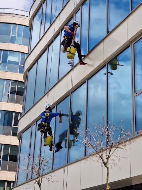 Two professional window cleaners wearing safety harnesses and helmets are performing surface cleaning on the large glass windows of a modern, multi-story office building in Knightsbridge. One cleaner, positioned higher up, is using a squeegee and cloth to wipe the reflective glass panes, while the other below is spraying cleaning solution onto the windows. The building features a curved façade with expansive glass panels, allowing natural light into the interior spaces. The scene is set during daylight with a clear blue sky reflected in the windows. This image highlights the expert commercial cleaning services offered by Carpet Cleaners Knightsbridge, focusing on surface sanitation and window maintenance within a professional environment.